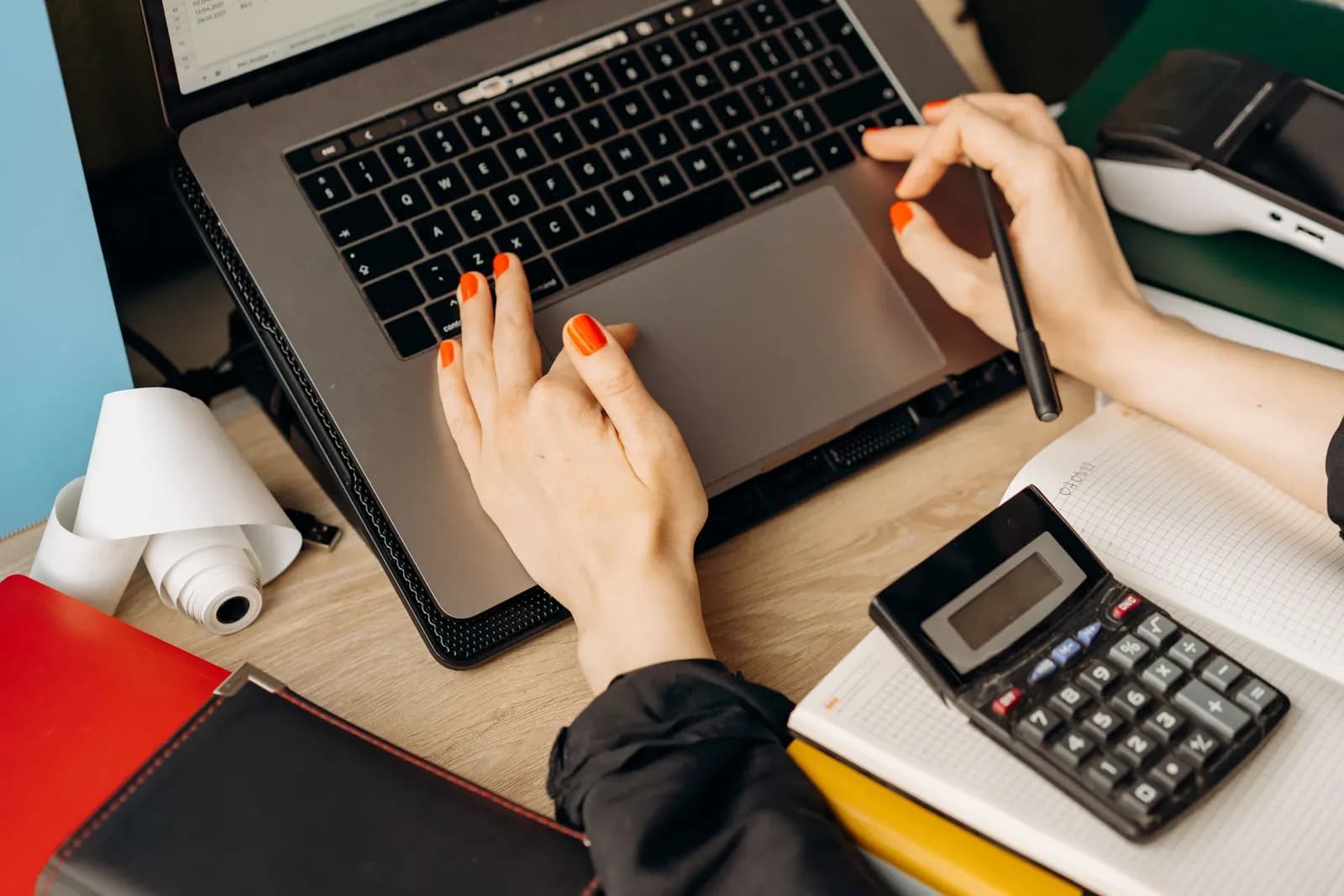 Hands working on a laptop beside a calculator, notebook and folders on a desk