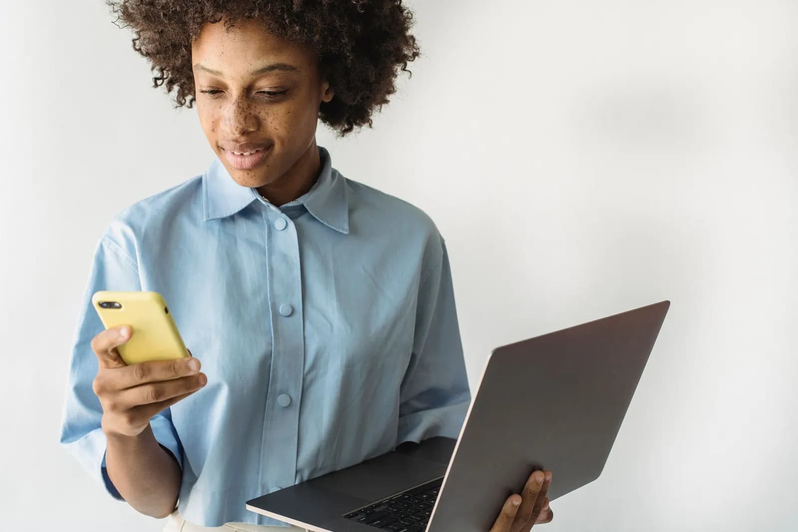 Woman looking at her smartphone while holding an open laptop