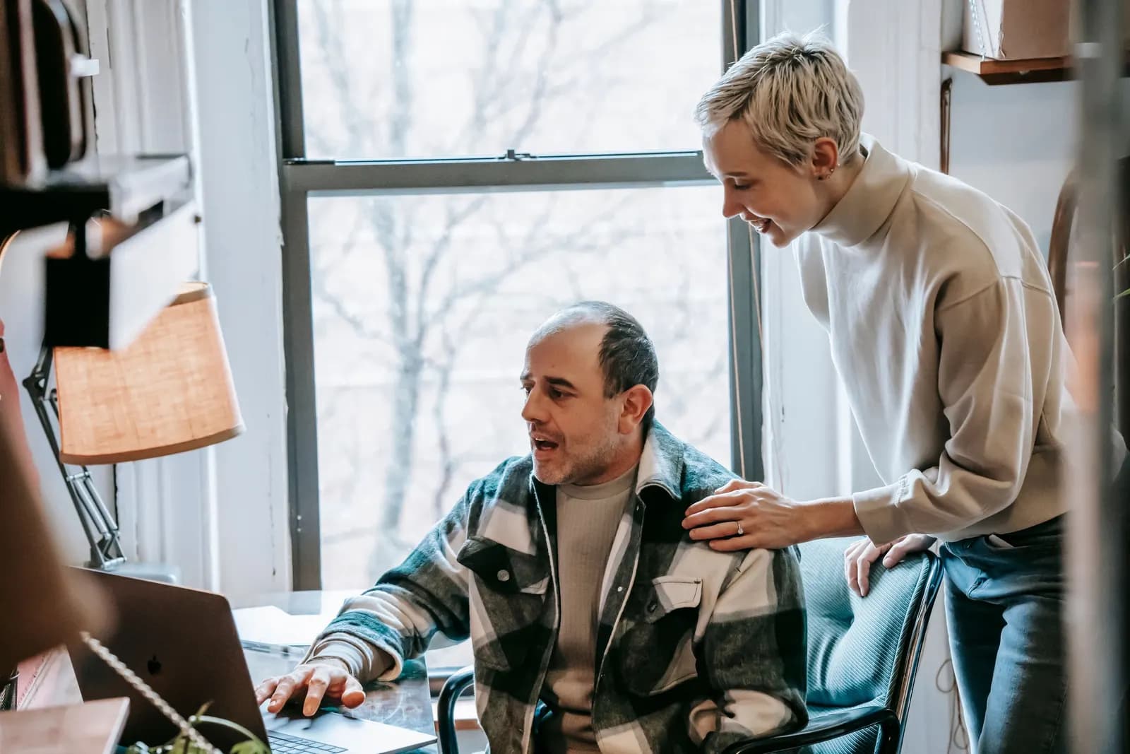 Woman leaning over to help a seated colleague with his laptop at a desk by a window